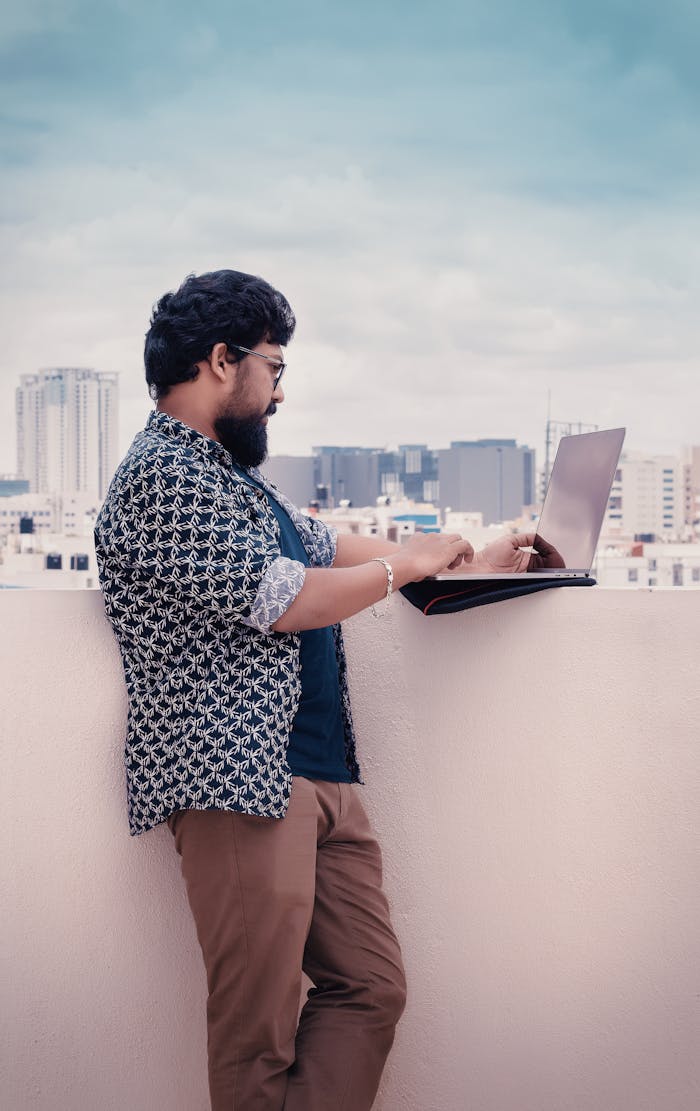 Man working on laptop on a rooftop with Bengaluru city skyline in the background.