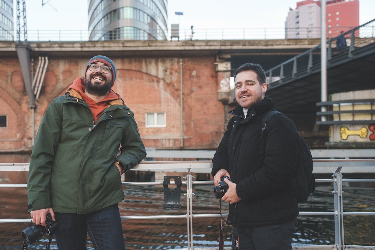 social-feed-img-04 Two friends with cameras enjoying photography near a river in an urban environment.